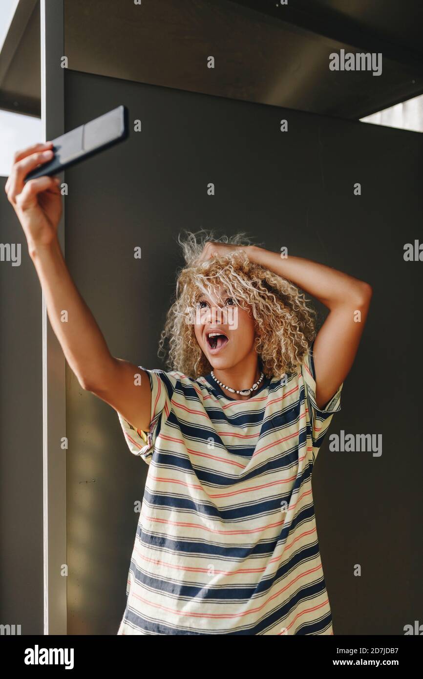 Donna che prende selfie attraverso smartphone mentre fa i volti contro parete metallica Foto Stock