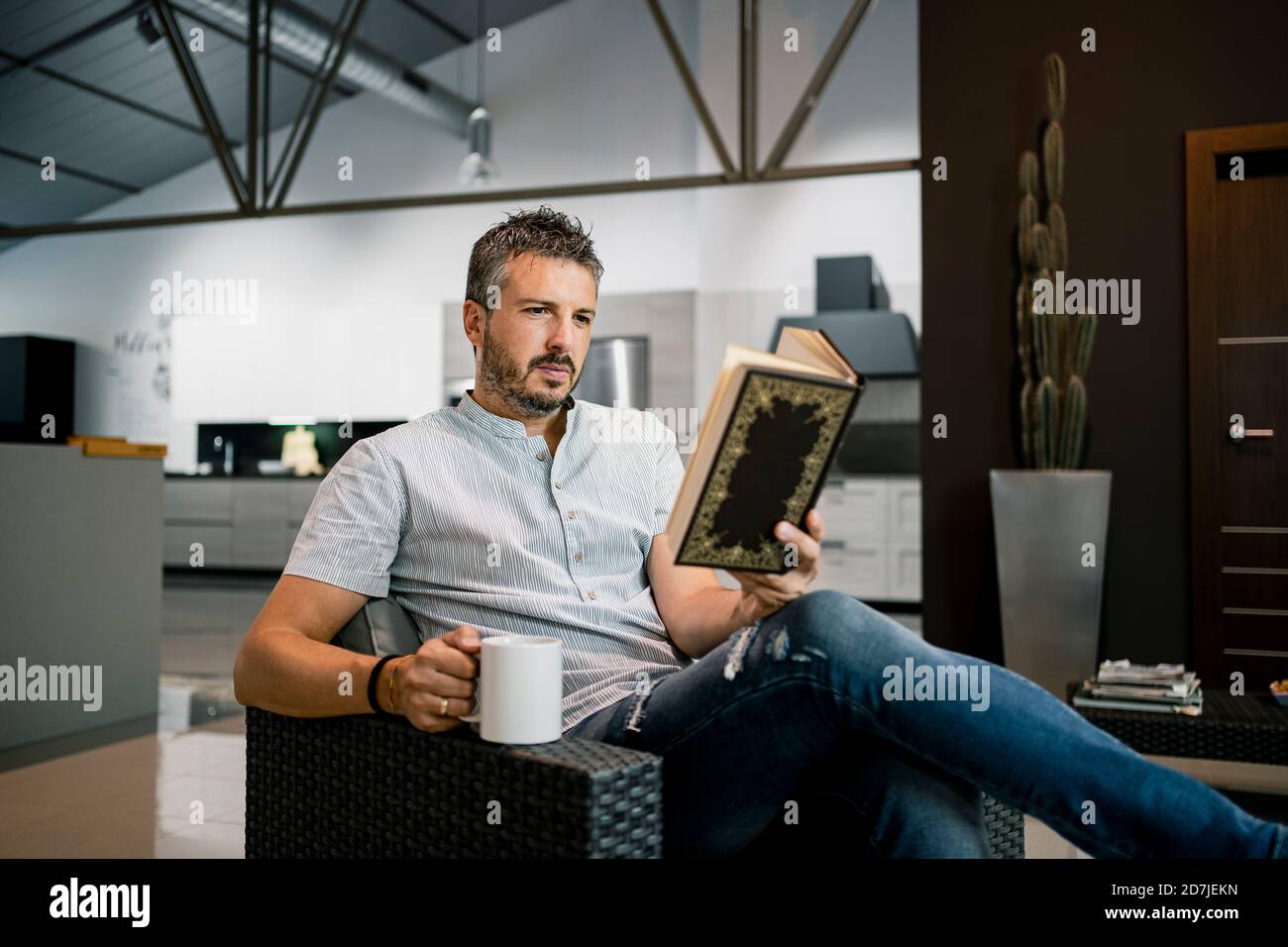 Rilassante libro di lettura freelance maschile mentre si siede con la tazza di caffè sulla sedia in ufficio Foto Stock