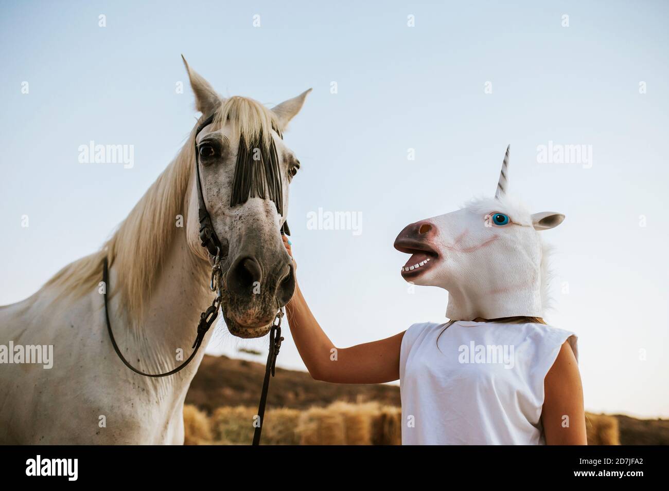 Giovane donna che indossa una maschera unicorno stracciando delicatamente il cavallo bianco Foto Stock