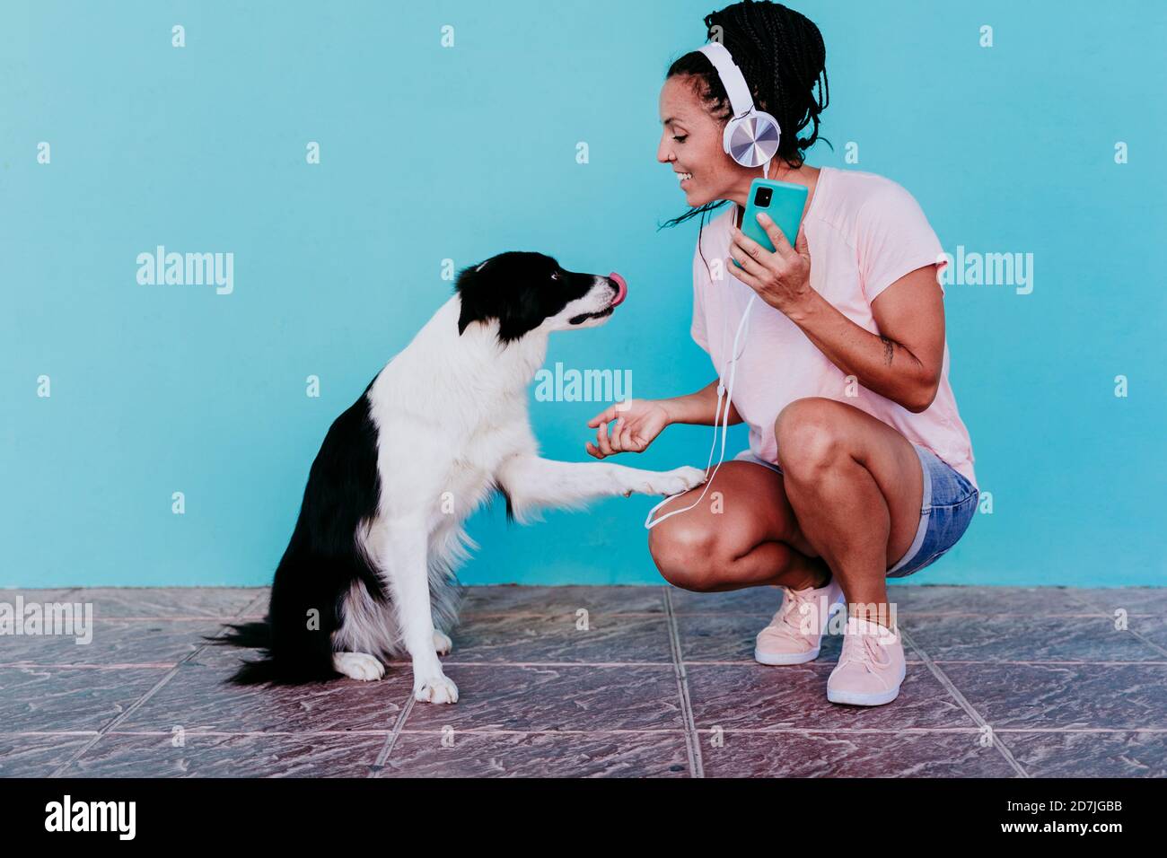 Donna sorridente che ascolta la musica attraverso le cuffie mentre si scuote Border Collie cane contro muro turchese Foto Stock