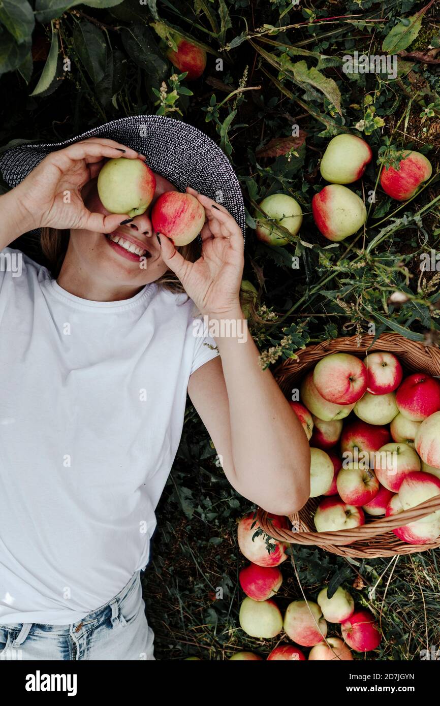 Donna sorridente che tiene le mele davanti agli occhi mentre si sdraiava a terra a frutteto Foto Stock