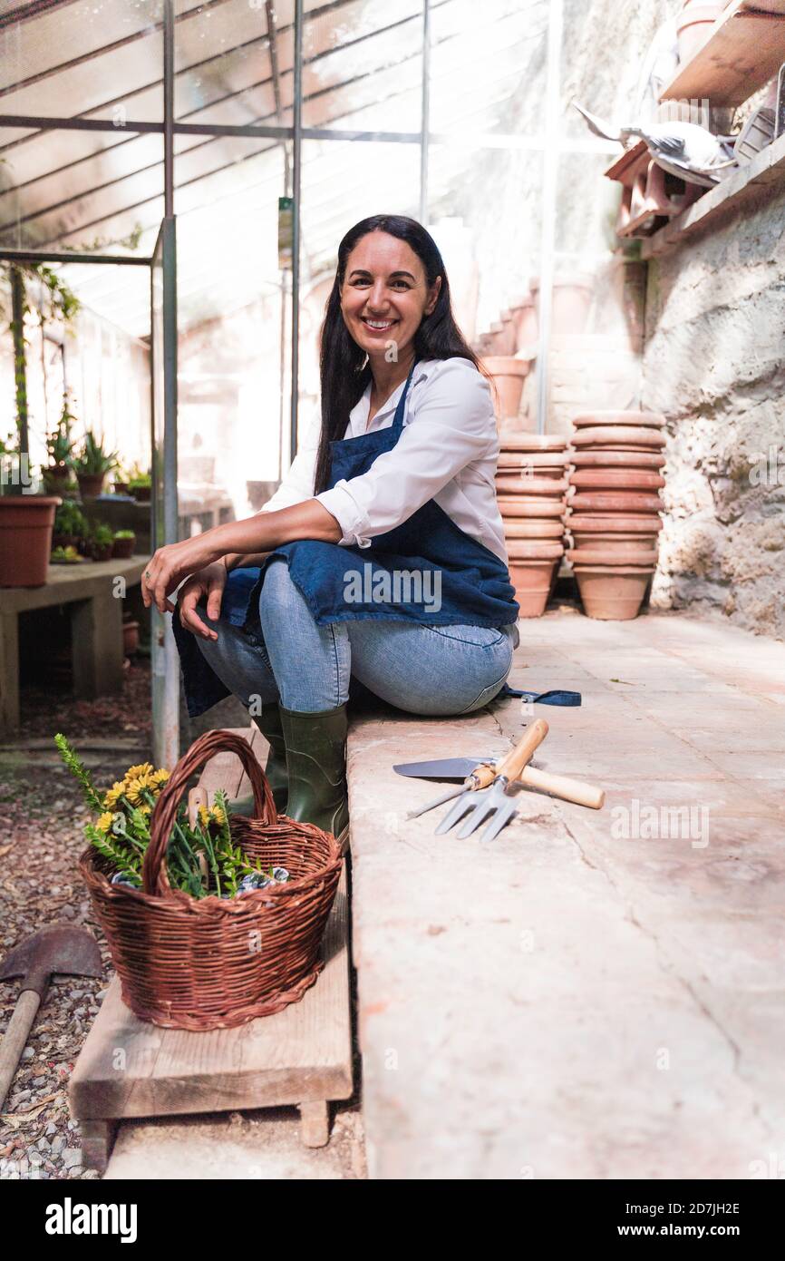 Donna sorridente seduta in capanna da giardino con cesto e giardinaggio attrezzatura in giornata di sole Foto Stock