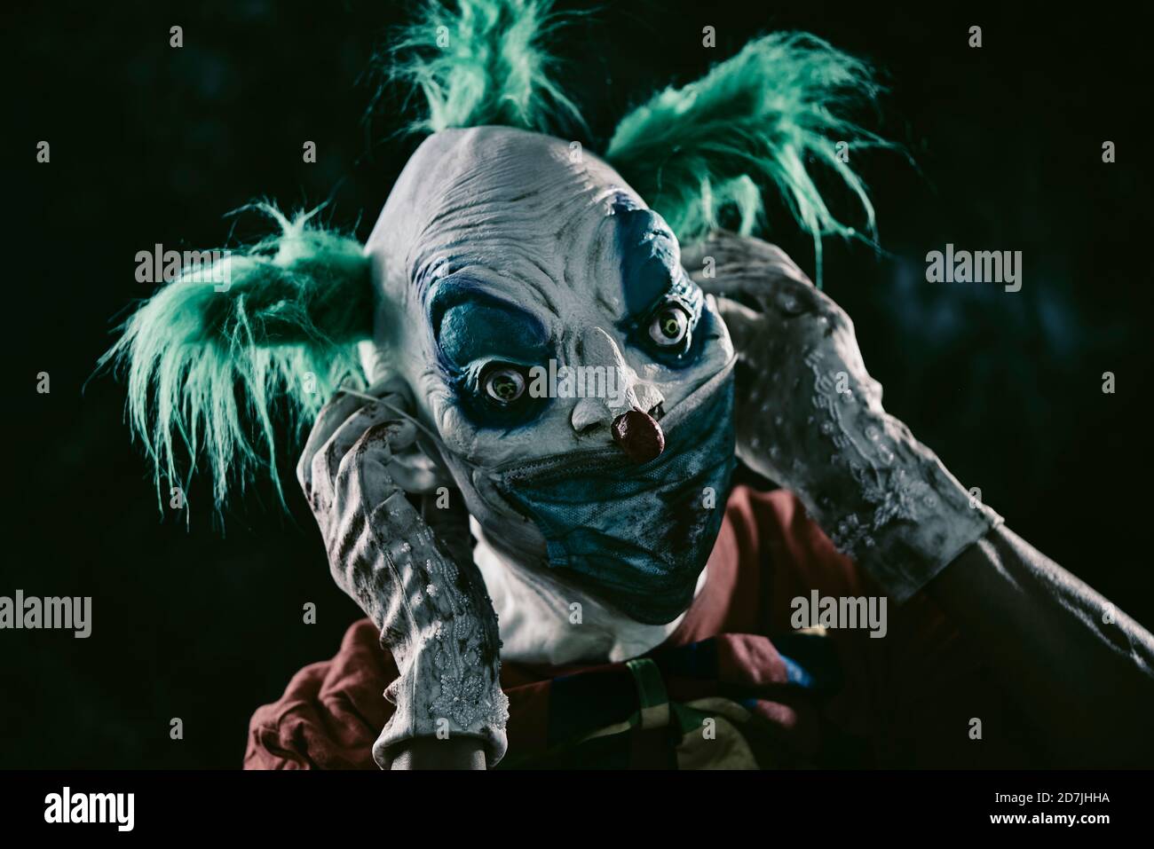 closeup di un clown male inquietante, con i capelli verdi, indossando un costume rosso sporco, una cravatta di arco sporca, e guanti sporchi e sanguinosi, indossando o prendendo Foto Stock