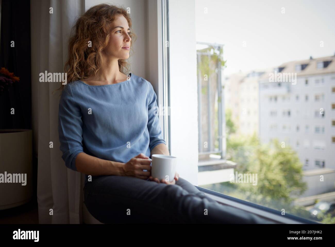 Donna con tazza di caffè che guarda attraverso la finestra mentre si siede  davanzale a casa Foto stock - Alamy