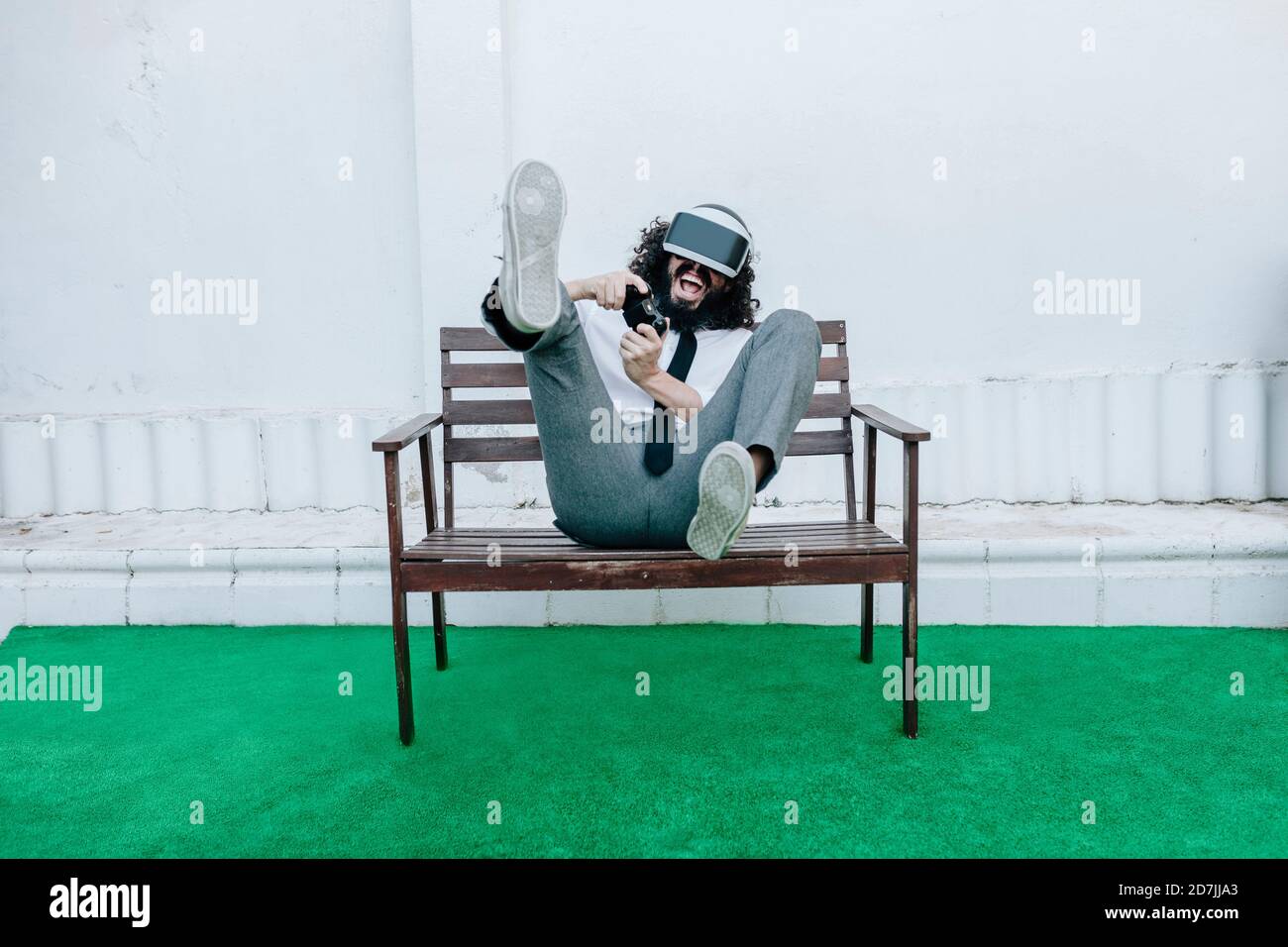 Uomo giocoso che gioca video gioco usando occhiali di realtà virtuale mentre seduto sulla panchina sul cortile Foto Stock