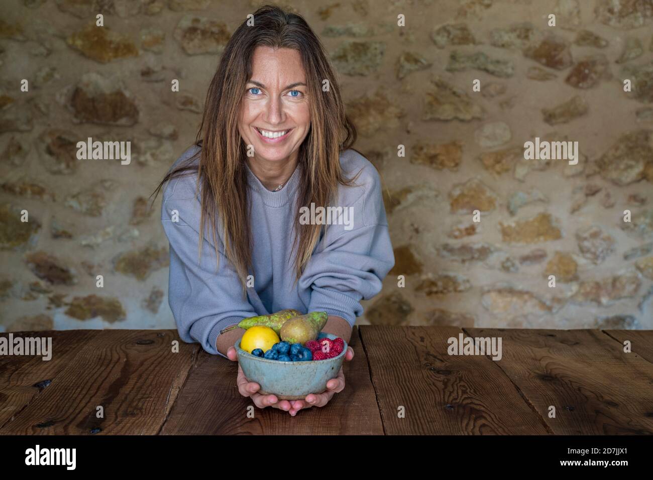 Sorridente nutrizionista femminile con vari frutti in ciotola seduta a. tavolo contro muro di pietra Foto Stock