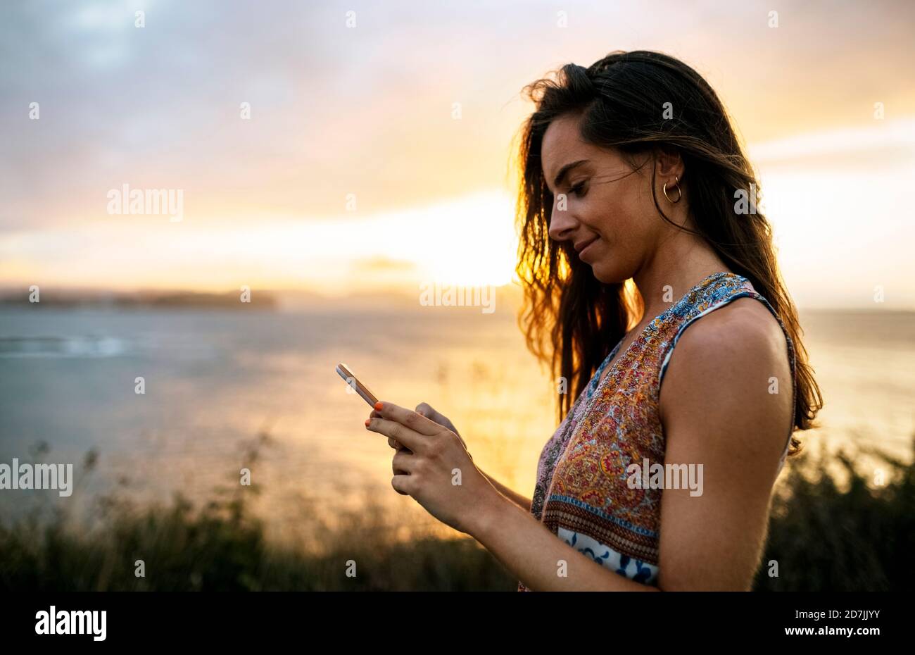 Sorridente bella donna che usa lo smartphone sulla spiaggia contro il cielo durante il tramonto Foto Stock