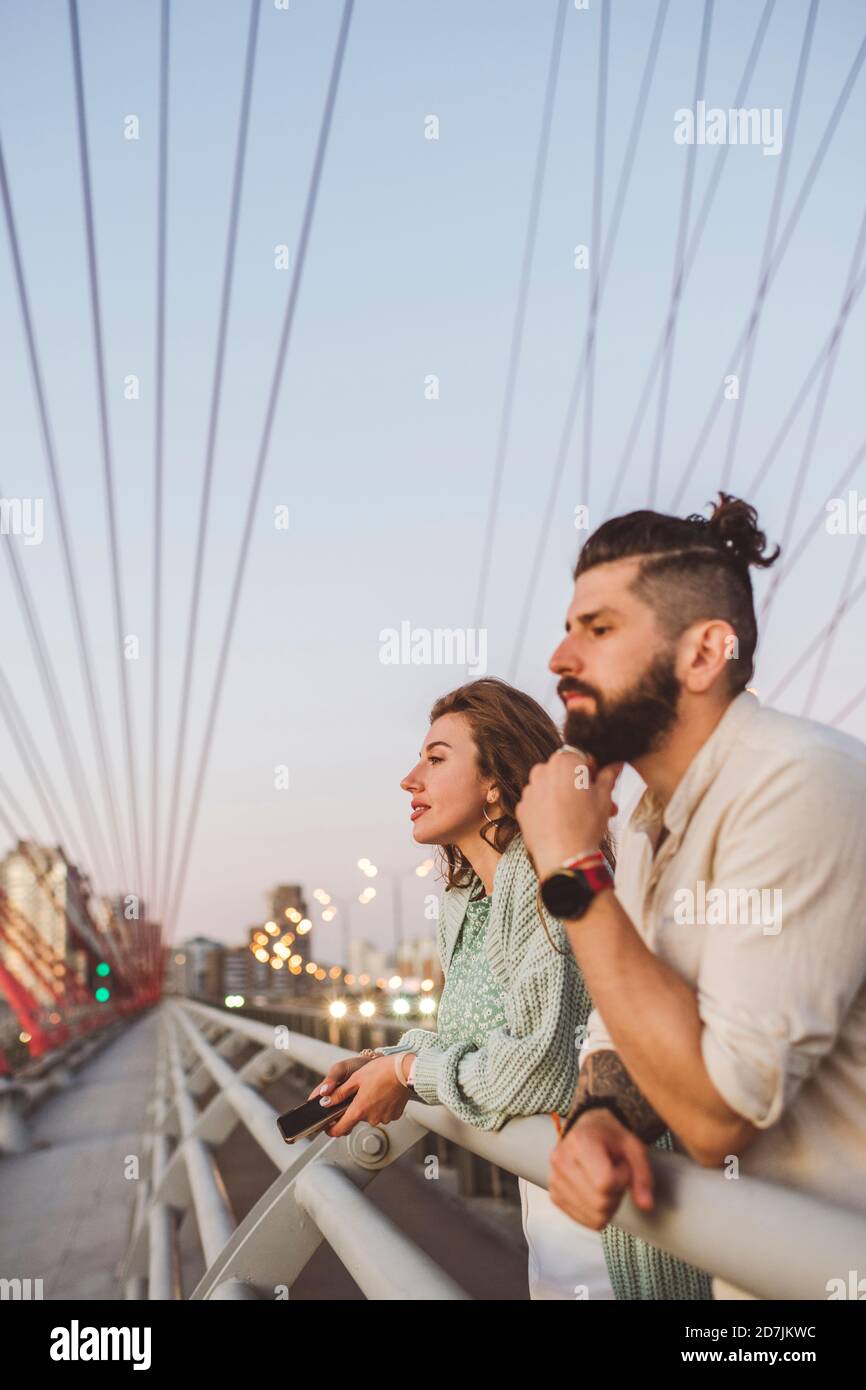 Coppia che guarda via mentre si appoggia sulla ringhiera al ponte dentro città durante il tramonto Foto Stock