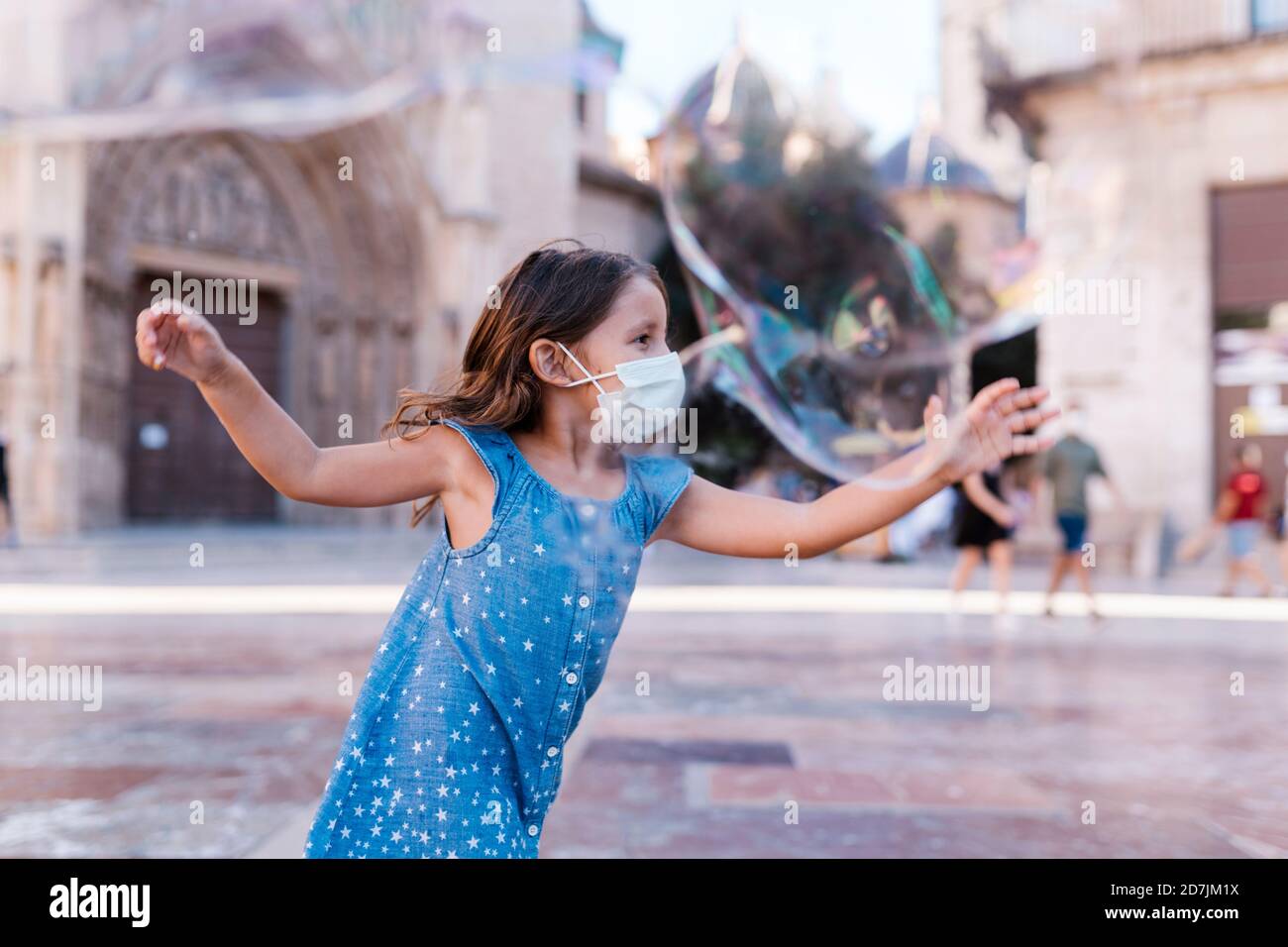 Ragazza spensierata indossando maschera che corre su strada in città giocando con bolla di sapone Foto Stock