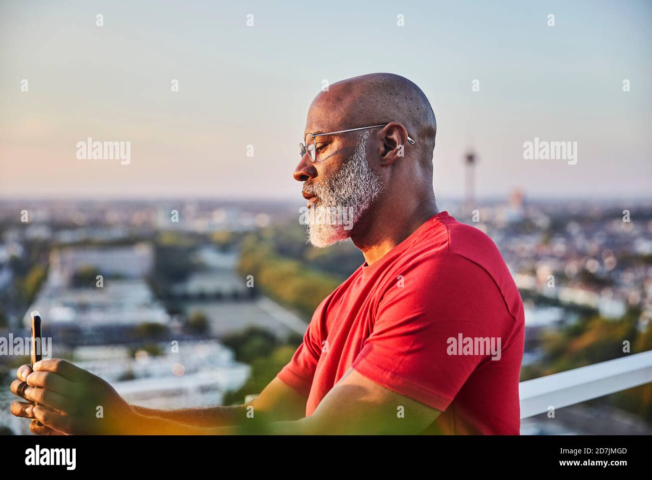Uomo calvo con barba bianca che usa lo smartphone per fotografare tramonto sulla città dalla terrazza Foto Stock