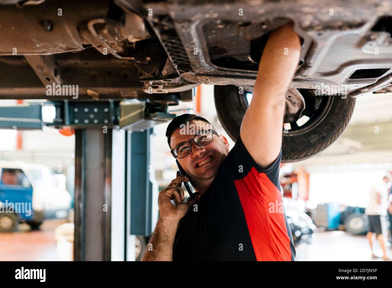 Meccanico che parla con il telefono cellulare durante la riparazione dell'auto in officina Foto Stock