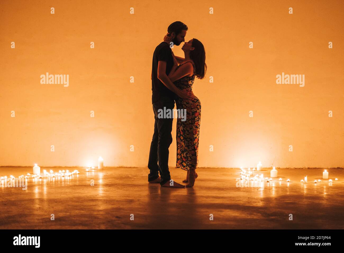 Una coppia romantica che si bacia e si abbraccia mentre si trova in mezzo a candele accese contro il muro a casa Foto Stock
