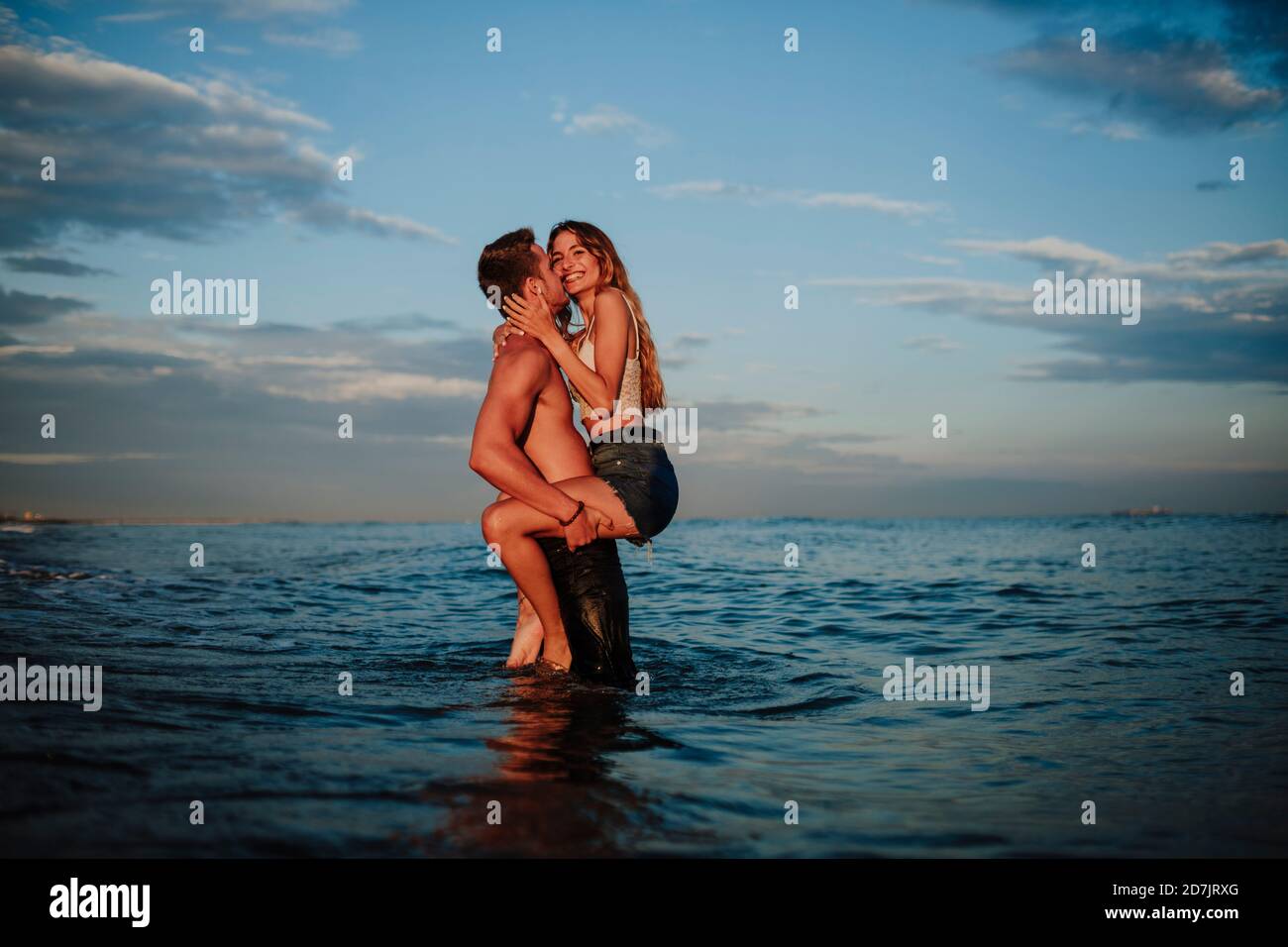 Uomo baciando e portando donna mentre si è in piedi in acqua durante tramonto Foto Stock