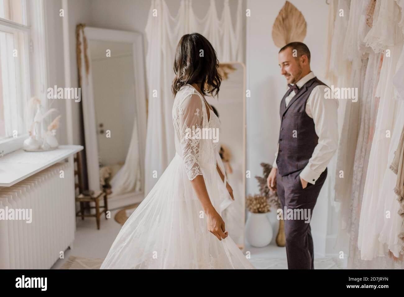 Giovane sposa che guarda nello specchio mentre lo sposo sta in piedi con le mani in tasca presso il negozio di abiti da sposa Foto Stock