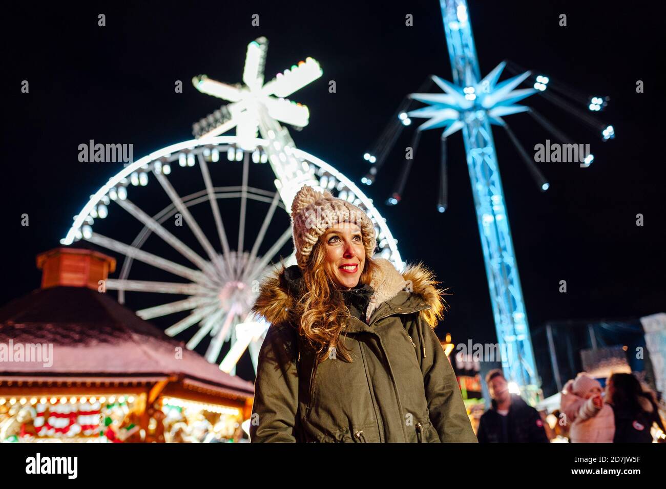 Donna sorridente che guarda via mentre si sta in piedi contro le giostre del parco divertimenti Di notte durante Natale Foto Stock