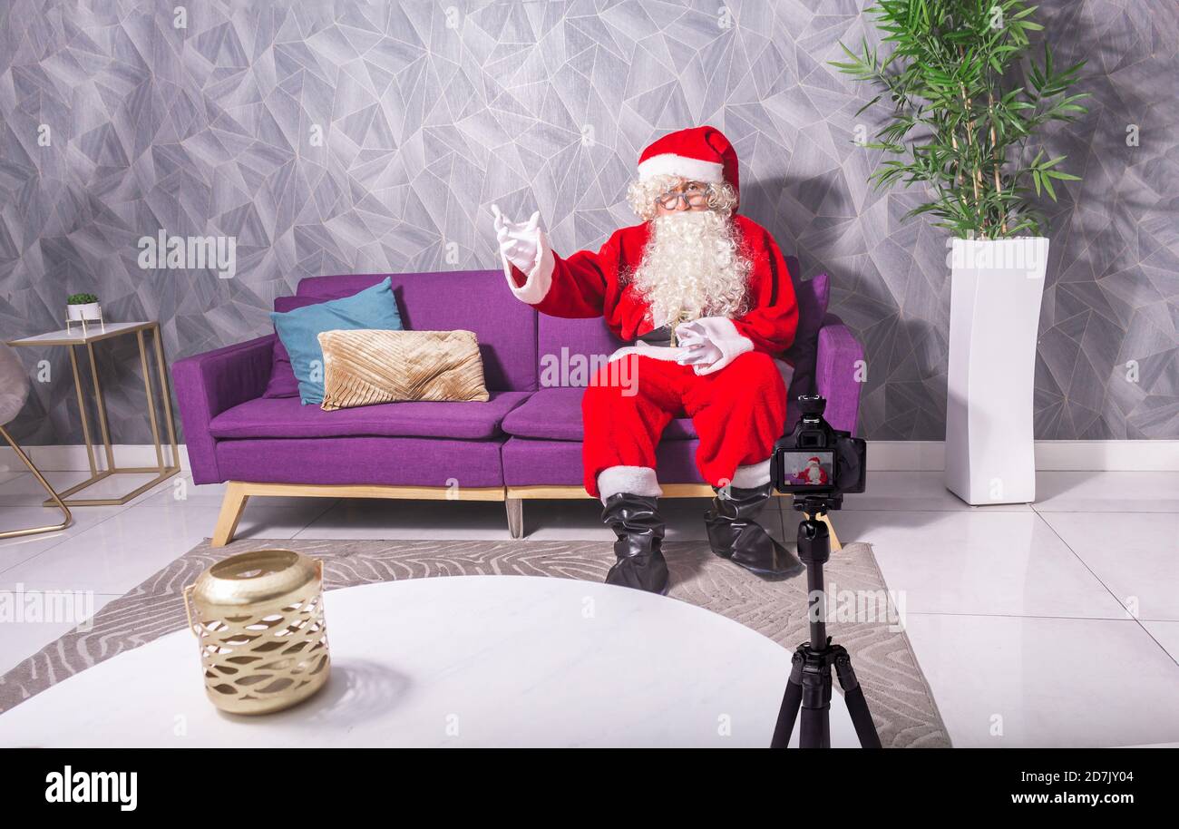 Babbo Natale a Natale registrando un video con una fotocamera dslr seduta su un divano in un ambiente moderno. Guardare sulla fotocamera in un tutorial Foto Stock