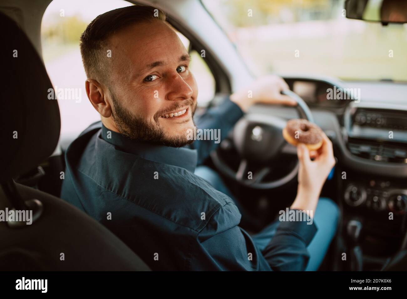 Un ritratto di un giovane uomo d'affari caucasico sorridente seduto in macchina e che tiene in mano una deliziosa ciambella di cioccolato. Un pasto dolce in trasporto Foto Stock