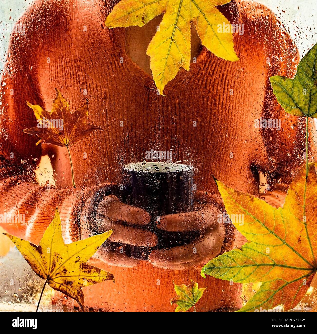 L'uomo in maglia calda arancione tiene una tazza di bevande calde e guarda attraverso la finestra con foglie d'autunno. Foto Stock