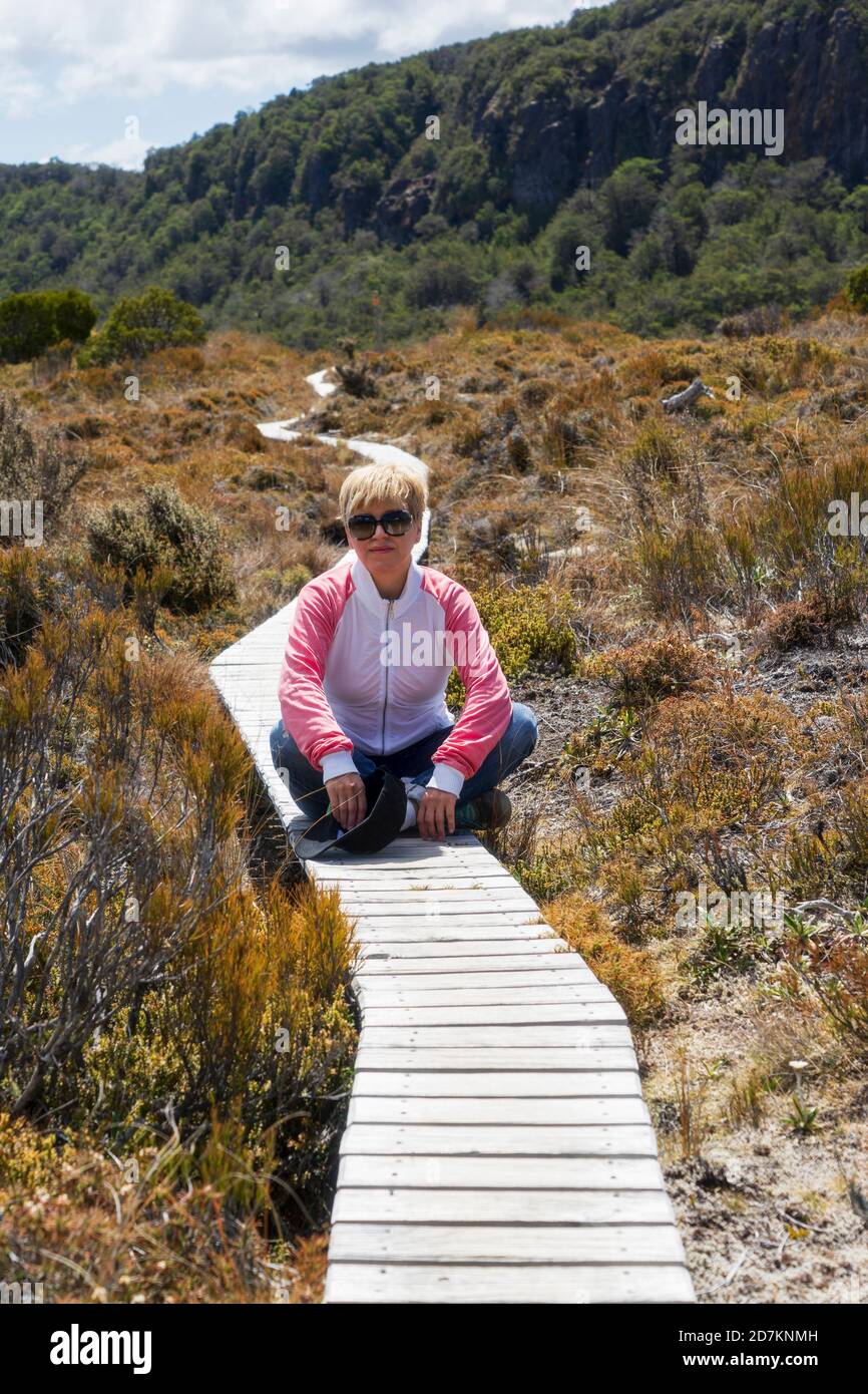 Donna in occhiali da sole seduta su sentiero turistico in legno che conduce a verdi colline. Percorso turistico al lago di sorpresa nella regione del vulcano Ruapehu. Foto Stock