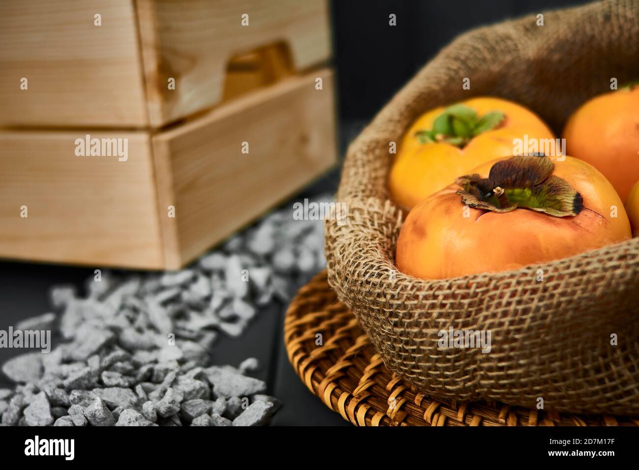 sacco di persimmons in primo piano su sfondo grigio, con pietre e una scatola di legno in tonalità arancio. Frutta autunnale Foto Stock