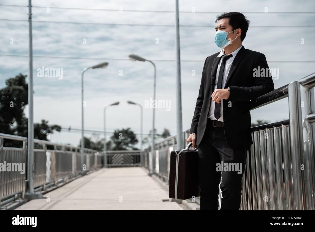 Disoccupato stressato giovane uomo d'affari asiatico indossare maschera portare una borsa di lavoro alla ricerca di lavoro in tuta copertura faccia con appoggiarsi contro una rotaia ponte. Non riuscito Foto Stock