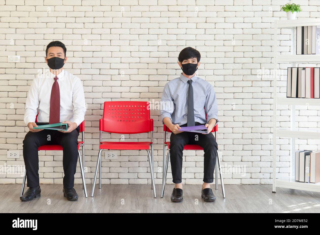 Due uomini asiatici si siedono in sedie di plastica rossa distanziate con Matt per coprire la bocca per evitare COVID-19 in attesa di un colloquio di lavoro. Foto Stock