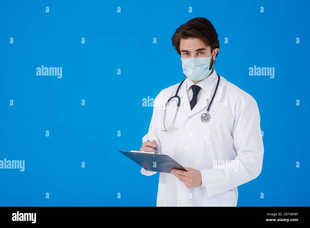 un giovane medico sopportato in un cappotto medico bianco e. una maschera su sfondo blu scrive il paziente reclami su un tablet con documenti e. Foto Stock