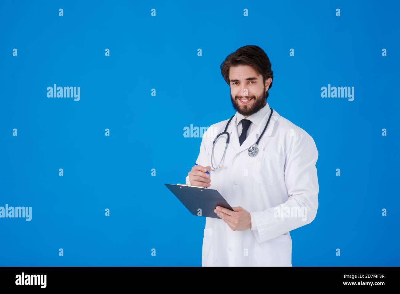 un giovane medico con la bearded in un cappotto medico bianco sopra uno sfondo blu scrive i reclami del paziente su un tablet con documenti e medici Foto Stock
