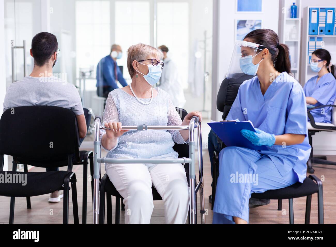 Donna anziana con telaio ambulante in sala d'attesa dell'ospedale con l'infermiere che indossa la maschera facciale contro la contaminazione dei coronavirus che discute di trattamento di malattia. Paziente e personale medico nell'area di attesa. Foto Stock