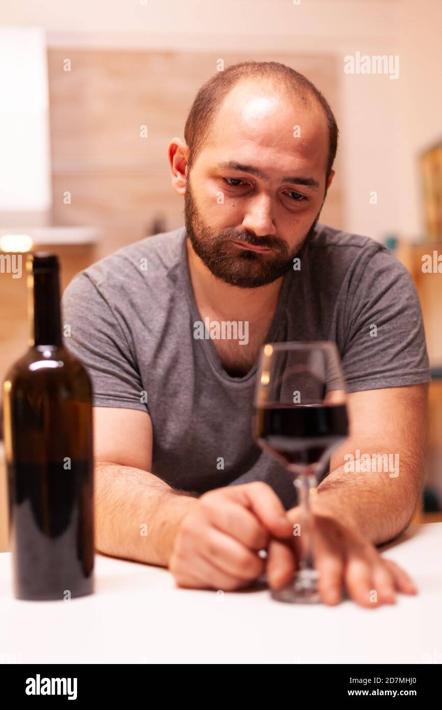L'uomo si siede depresso e solo con un bicchiere di vino rosso al tavolo da  cucina. La malattia della persona infelice e l'ansia si sentono esauriti  con problemi di alcolismo Foto stock -