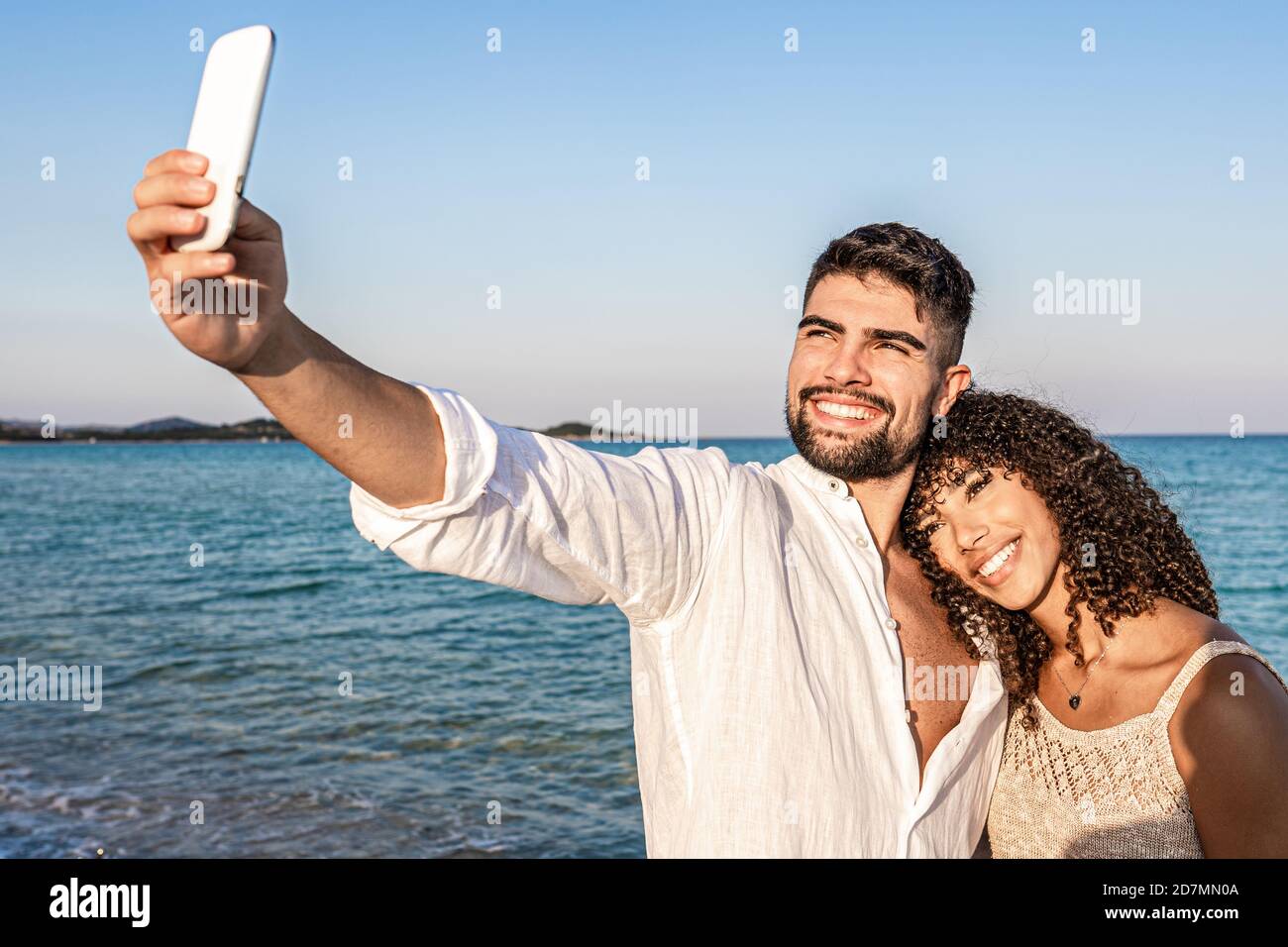 I giovani amanti della corsa mista si accoppiano al tramonto il mare fa un ritratto automatico con lo smartphone - Uomo caucasico che ha divertimento sparando ph Foto Stock