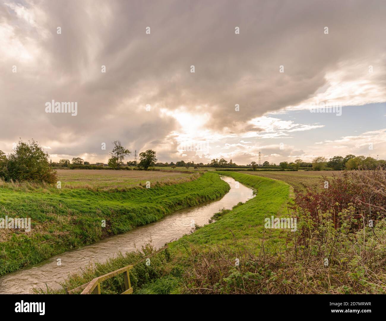 Un piccolo fiume curva tra due campi che sono stati arati dopo il raccolto. I piloni sono all'orizzonte e una nuvola pesante è in testa. Foto Stock