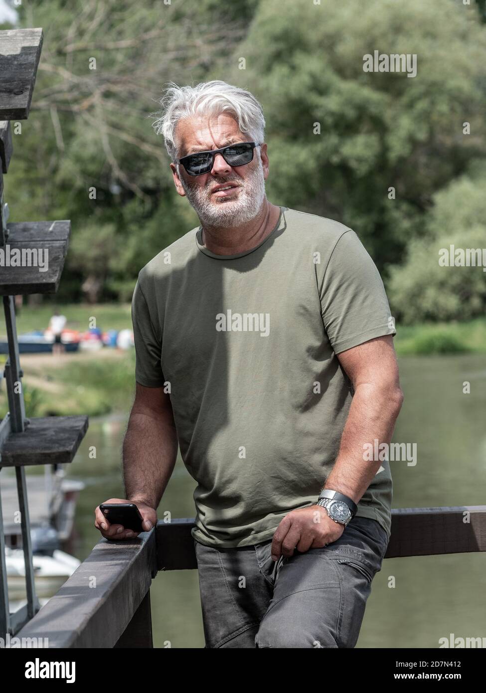 Bell'uomo anziano con capelli grigi, barba, T-shirt verde e occhiali da sole in piedi all'aperto. Sfondo sfocato. Foto Stock