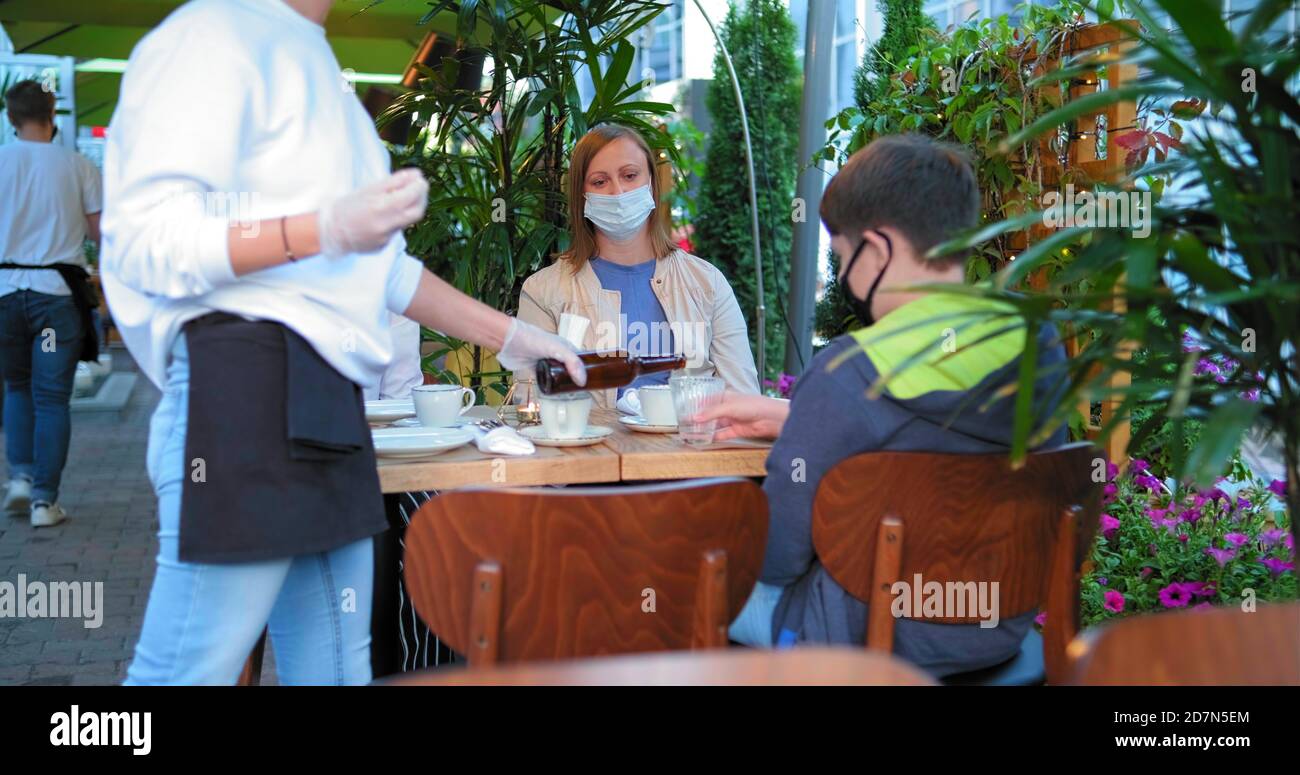 la cameriera in guanti sterili versa il succo in un bicchiere per bambini e. mamma con amico in maschere di faccia sedersi a legno marrone tavolo sulla terrazza Foto Stock