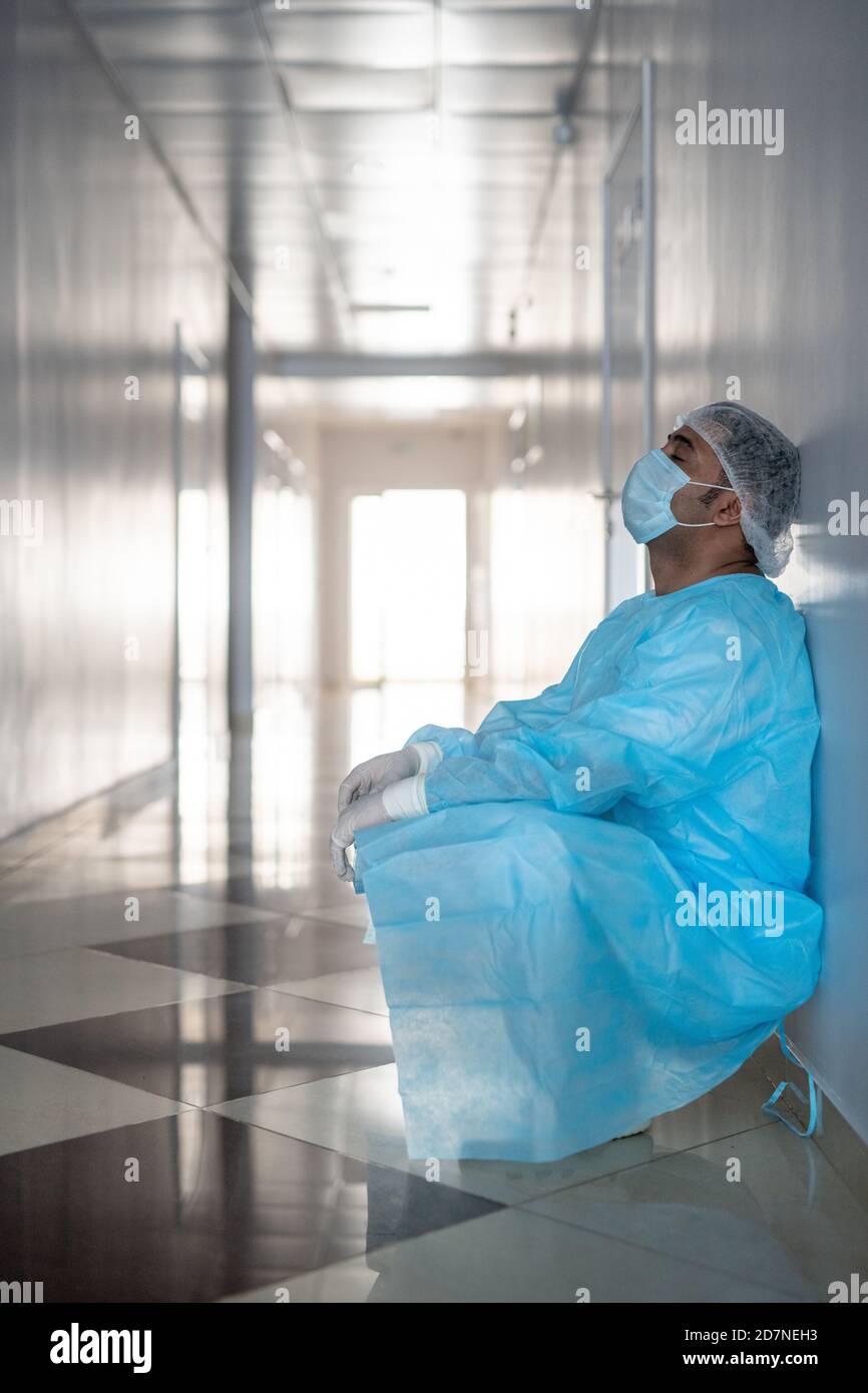 Chirurgo maschile stanco professionale in indumenti da lavoro protettivi appoggiati alla parete Foto Stock