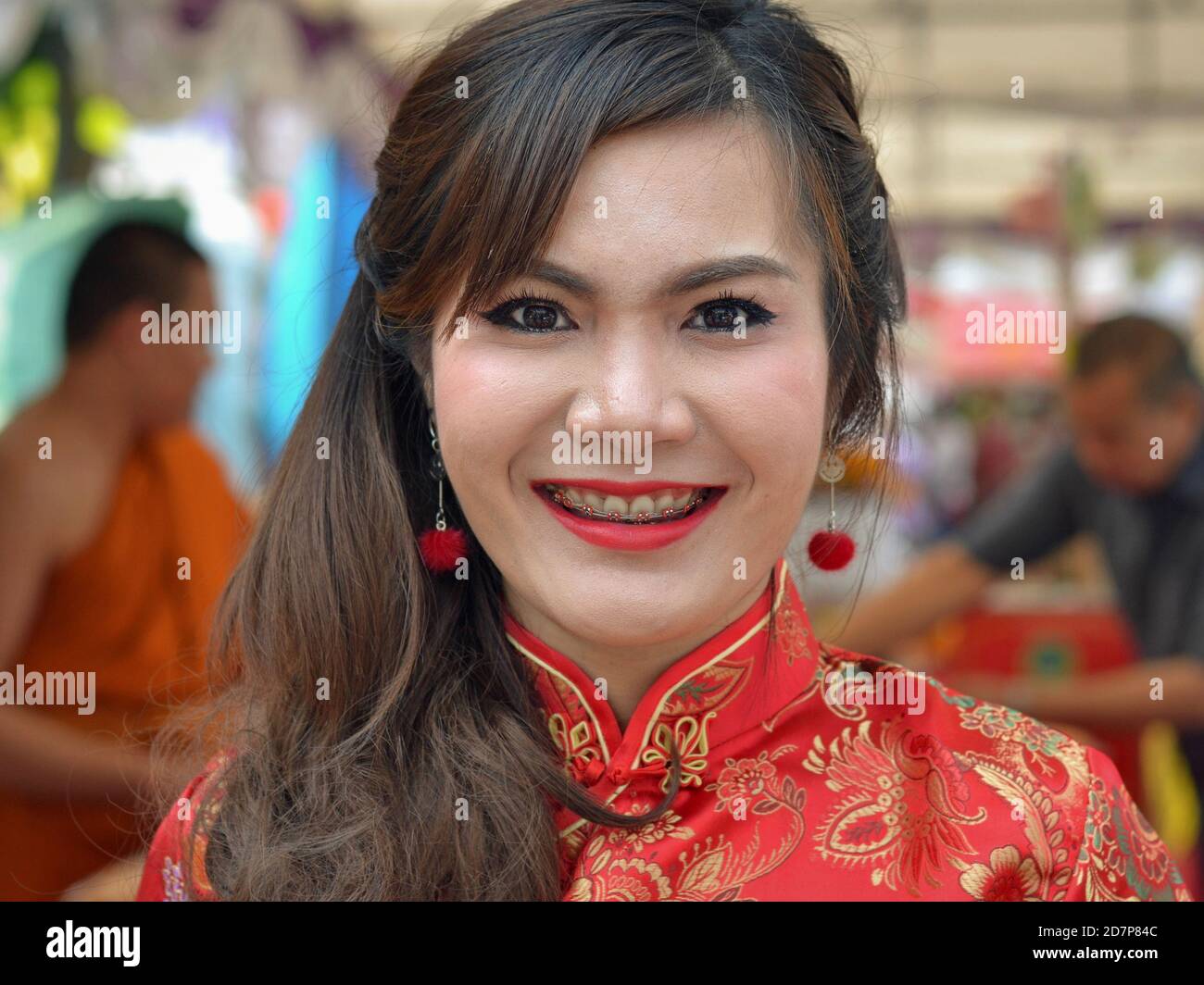 Giovane donna thailandese con bretelle dentali rosse, rossetto rosso e abito di seta cinese sorride per la macchina fotografica all'interno di un tempio buddista durante il Capodanno cinese. Foto Stock