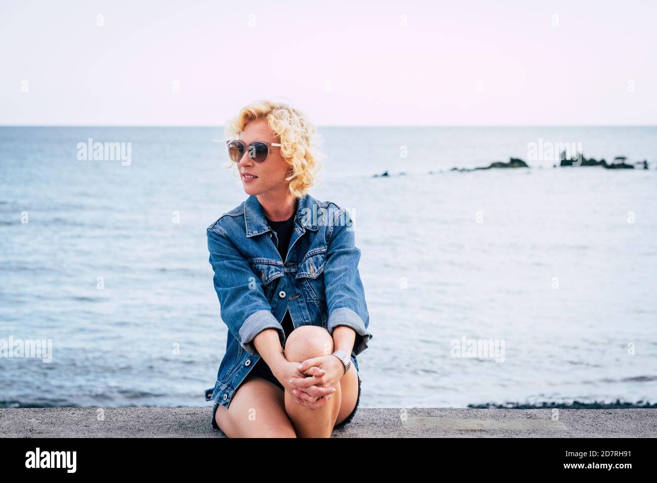 Ritratto adulto di mezza età di bella donna bionda singola seduta con oceano e cielo sullo sfondo - persone all'aperto attività per il tempo libero rilassarsi Foto Stock