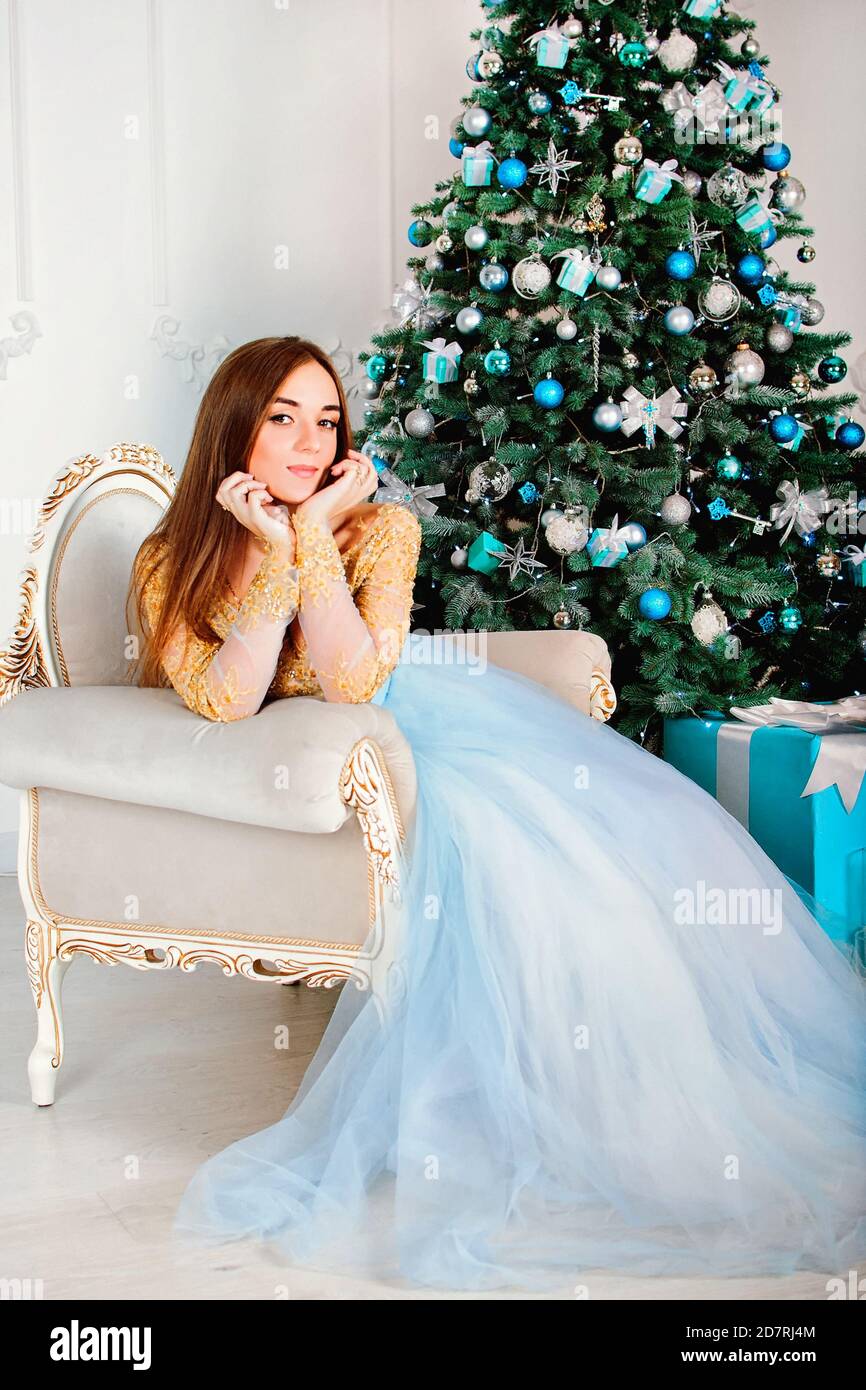 Donna giovane e felice in abito da festa lungo seduto vicino all'albero di Natale con i giocattoli di Natale in studio di festa. Cartolina invernale. Cartolina di Natale. Foto Stock