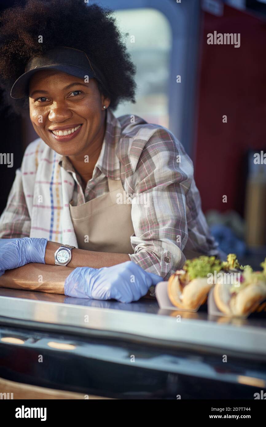 ritratto di una giovane donna afro-americana in fast food, guardando la macchina fotografica, sorridendo, appoggiata con le mani incrociate, indossando guanti con rea Foto Stock