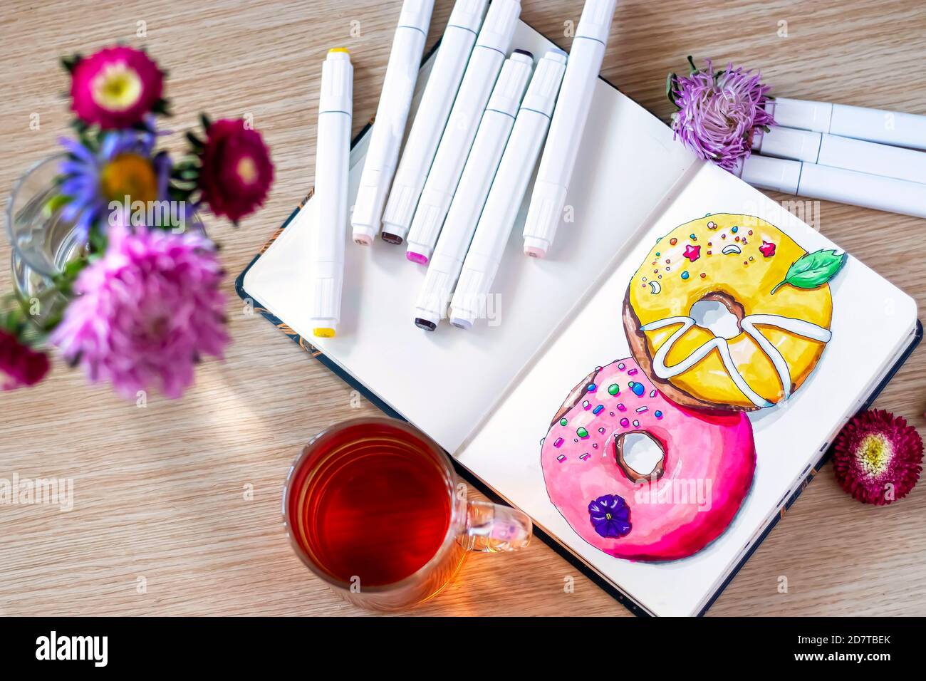 Disegno di ciambelle nel notebook. Il tutto posto su un tavolo di legno con una tazza trasparente di tè, taccuino, pennarelli e fiori. Hobby. Ciambelle da disegno. Vista dall'alto. Messa a fuoco morbida Foto Stock