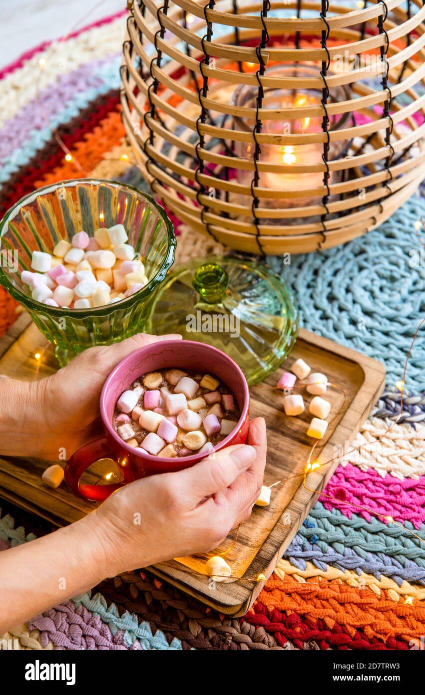 Mani che tengono una tazza rossa di cacao con marshmallows di colore pastello, lanterna di filo e bambù con candela su colorato tappeto di uncinetto da T-shirt ridisposte. Foto Stock