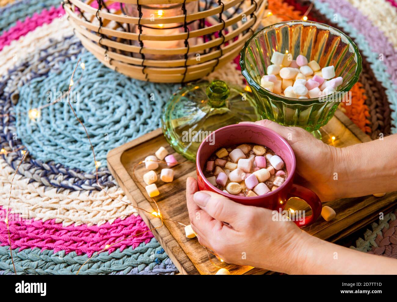 Mani che tengono una tazza rossa di cacao con marshmallows di colore pastello, lanterna di filo e bambù con candela su colorato tappeto di uncinetto da T-shirt ridisposte. Foto Stock