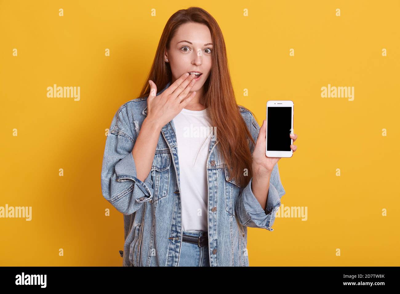 Primo piano ritratto di giovane bella donna scioccata abiti casual stile, tenendo telefono con schermo vuoto e tiene il palmo sulla sua bocca, guardando camer Foto Stock