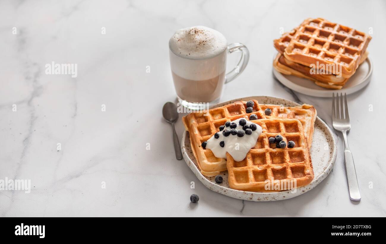 Deliziosi waffle belgi fatti in casa con yogurt greco, mirtilli e cappuccino su sfondo di marmo bianco. Colazione perfetta con spazio per testi o disegni Foto Stock