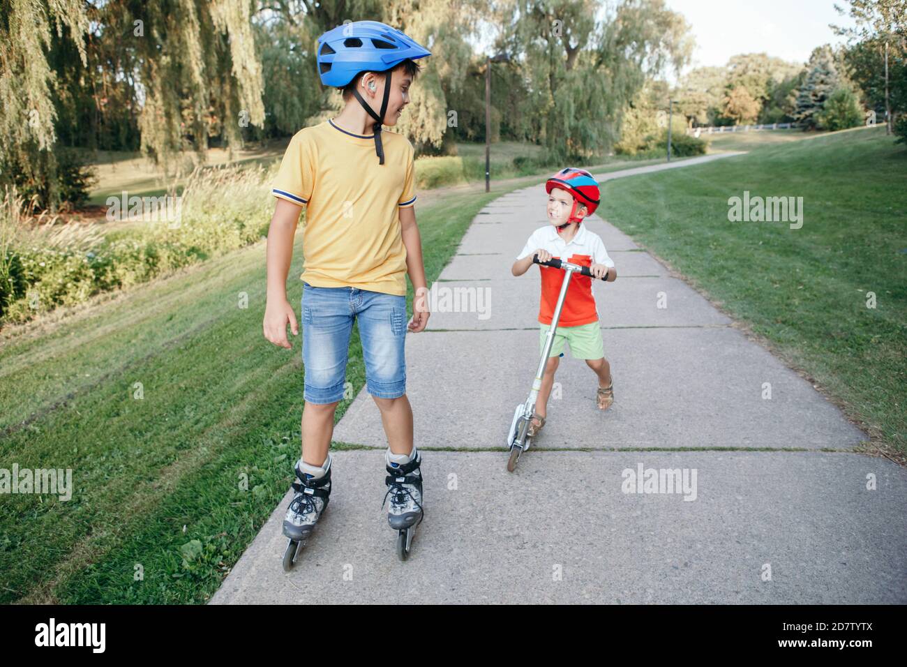 Ragazzi caucasici fratelli in caschi a cavallo pattini a rotelle e scooter su strada nel parco. Sport stagionale all'aperto per bambini. Vita sana dell'infanzia Foto Stock