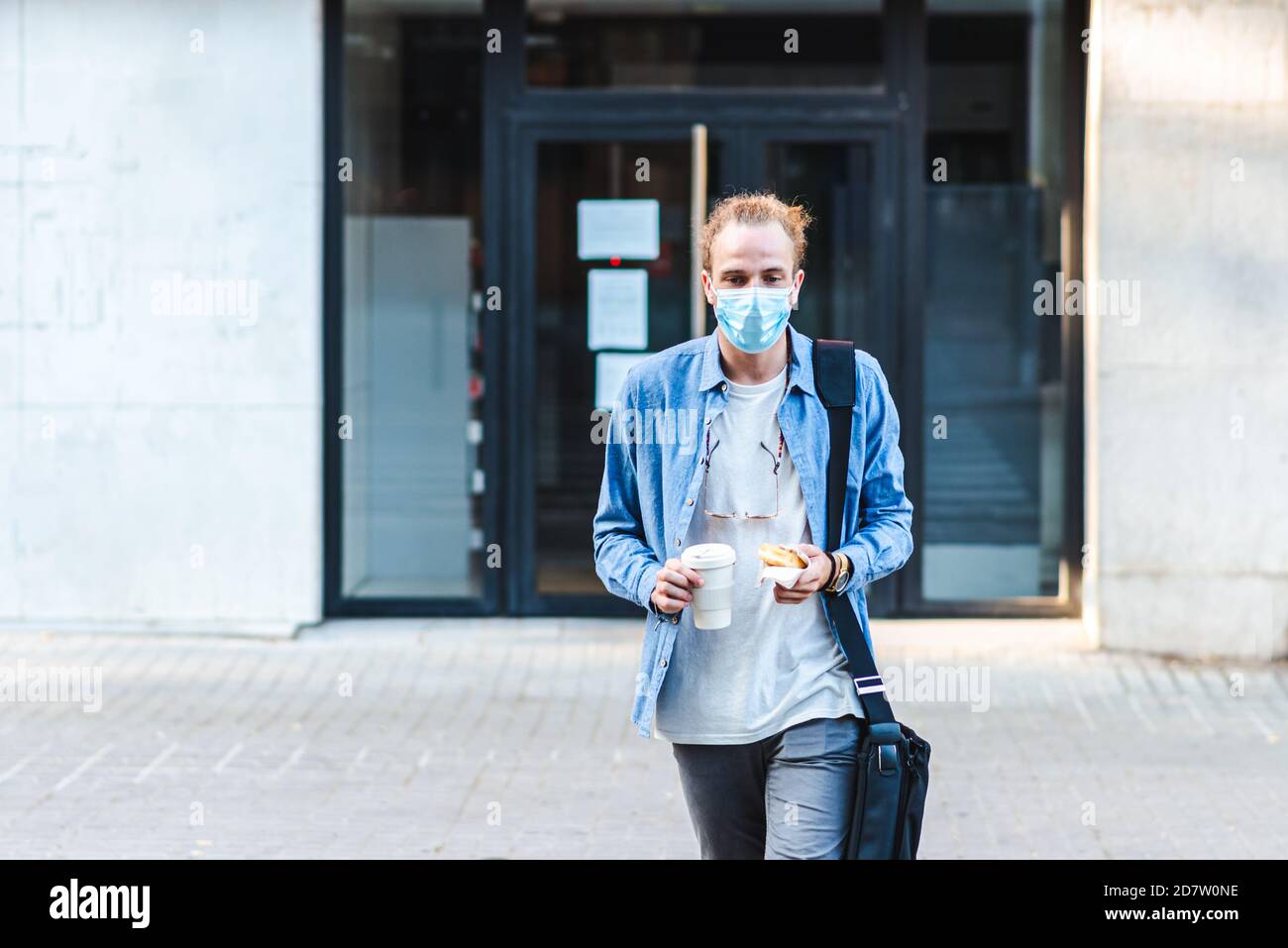 Giovane caucasico che indossa casual e con una maschera chirurgica tenendo una ciambella e un thermos di traversata del caffè da asporto la strada di una città Foto Stock