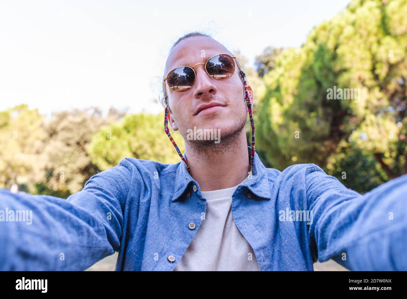 giovane uomo caucasico che indossa occhiali da sole a braccia aperte guardando a macchina fotografica con alberi sullo sfondo. concetto di auto-immagine Foto Stock