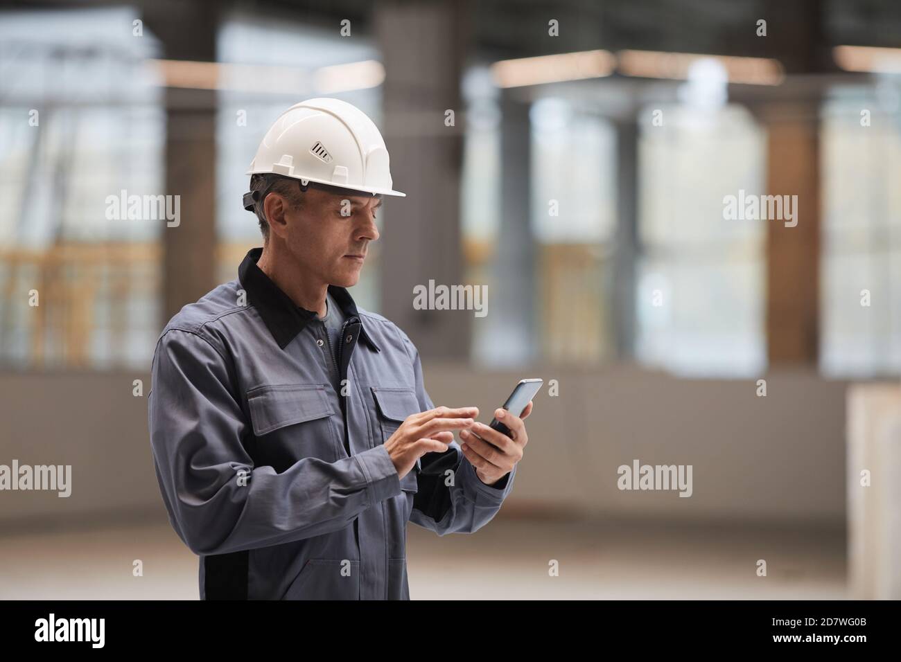 Vista laterale ritratto di un lavoratore maturo che utilizza uno smartphone mentre si trova in cantiere o in officina industriale, spazio di copia Foto Stock