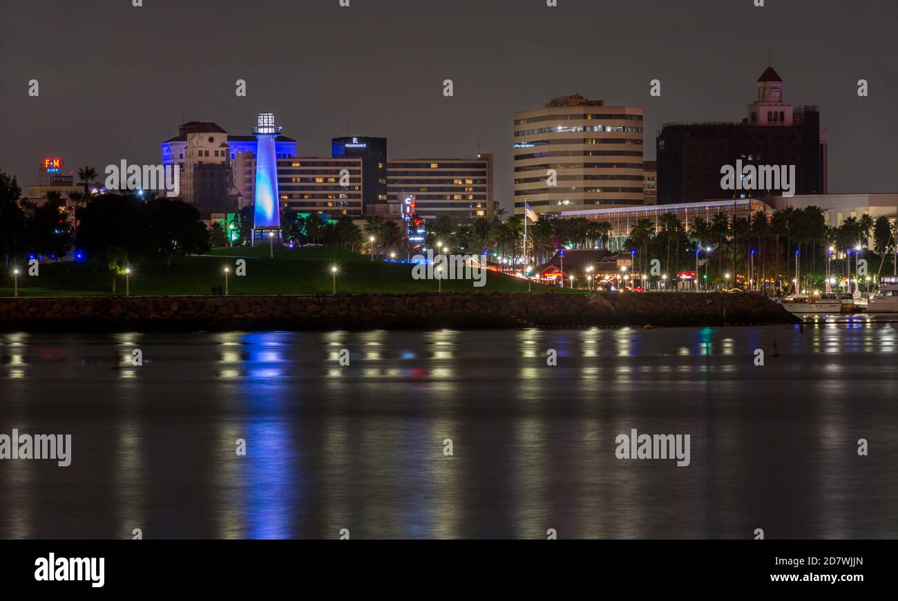 Foto notturna di un parco cittadino vicino al centro di Long Beach con vista sul Rainbow Harbour, con skyline e luci della città, oltre a un faro illuminato. Foto Stock