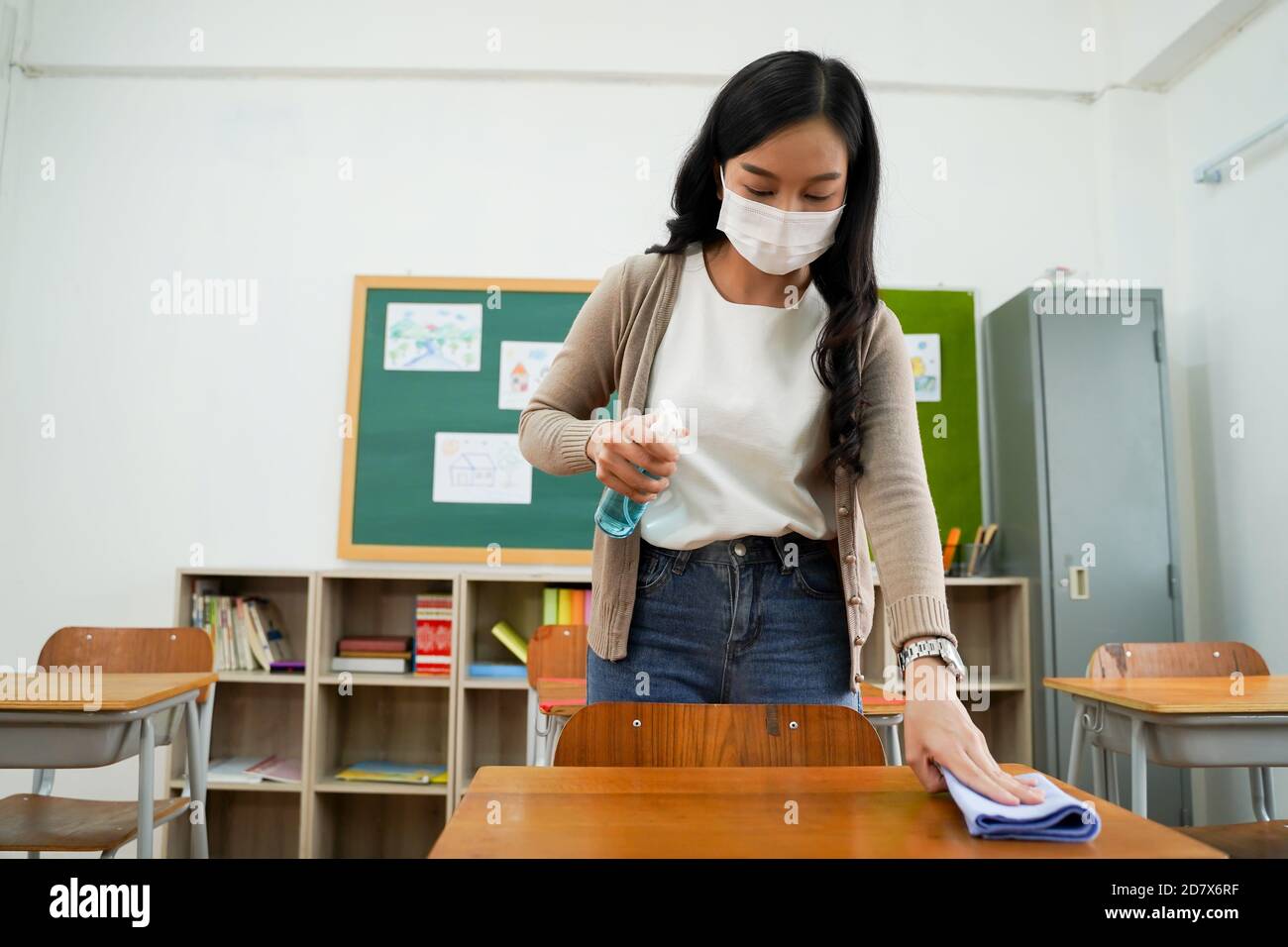 Giovane insegnante di sesso femminile che usa uno spray alcolico per  disinfettare le scrivanie degli studenti in classe. Donna asiatica in  maschera viso pulire i tavoli con disinfettante antisettico. Riaprire la  scuola